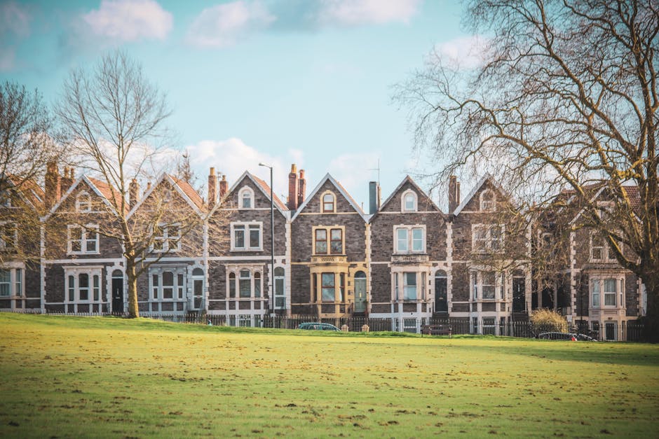 A row of Victorian-style terraced houses on Earls Court Road with dark brick facades, white window frames, and pitched roofs, set against a bright blue sky with some white clouds. The foreground features a well-maintained grassy area with some leafless trees, and the façade of each house includes bay windows, front doors, and chimneys. The scene is illuminated by natural daylight, highlighting the neat cleanliness and historic architectural details, reflecting the importance of surface cleaning and maintenance, as promoted by Carpet Cleaners Earls Court for residential hygiene and deep cleaning.