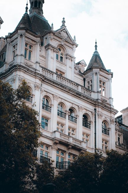 A historic Victorian-style building with ornate architectural details, multiple arched windows, decorative balconies with black wrought-iron railings, and pointed turrets topped with spires. The building's facade is light-colored stone or plaster, featuring intricate moldings and columns. Surrounding the structure are lush green trees, partially obscuring the lower floors, with soft natural light illuminating the scene. This elegant property exemplifies classic design, suitable for deep cleaning or surface sanitisation by Carpet Cleaners Earls Court, specialists in domestic cleaning services.