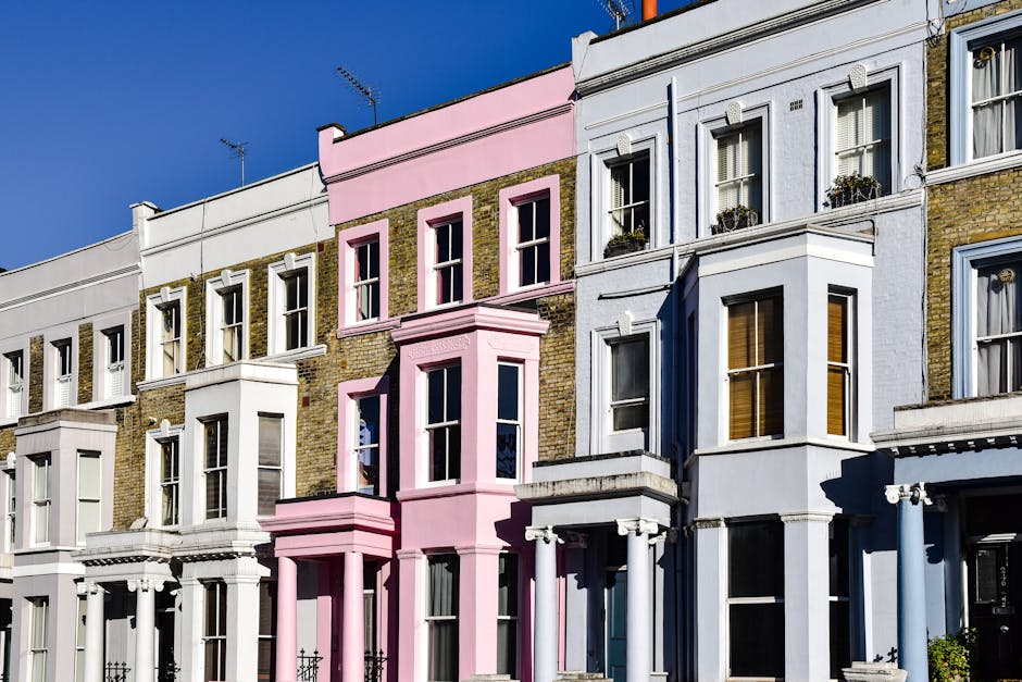 A row of multi-story residential buildings with facades in white, pastel pink, and light blue, featuring bay windows, decorative moldings, and small balconies with potted plants. The buildings have brickwork and painted surfaces, set under a clear blue sky with bright sunlight casting shadows, illustrating an urban street scene in Earls Court. The image represents well-maintained exteriors, emphasizing cleanliness and aesthetic decor, consistent with professional cleaning and maintenance standards, as highlighted by Carpet Cleaners Earls Court’s surface cleaning and deep cleaning services.