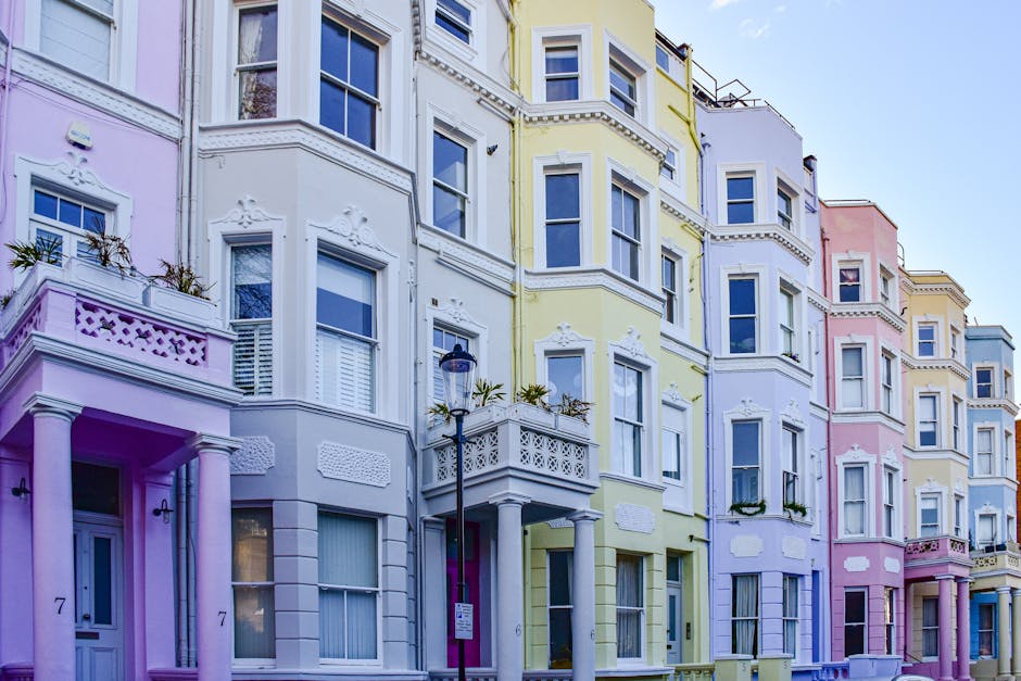 Exterior view of a row of multi-story residential buildings in Earls Court, painted in pastel shades of pink, yellow, lavender, and white, with ornate architectural details and decorative moldings around windows. The buildings feature large windows with white frames, some with shutters, and small balconies with intricate railing designs. A black street lamp is positioned along the sidewalk, which is visible at the base of the image. The scene is illuminated by natural daylight, highlighting the clean and well-maintained facades of the buildings, indicative of a tidy, urban residential area. Carpet Cleaners Earls Court specializes in surface and deep cleaning services, ensuring hygienic conditions for properties like these.