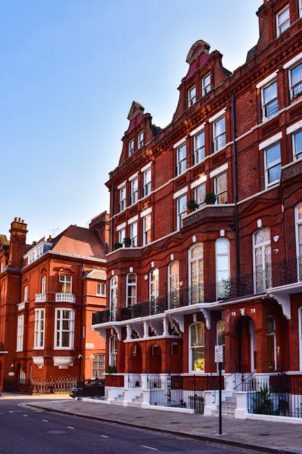 An urban street scene featuring a corner building with Victorian architectural details, including ornate white window frames and decorative brickwork, situated on a paved road with parked cars along t